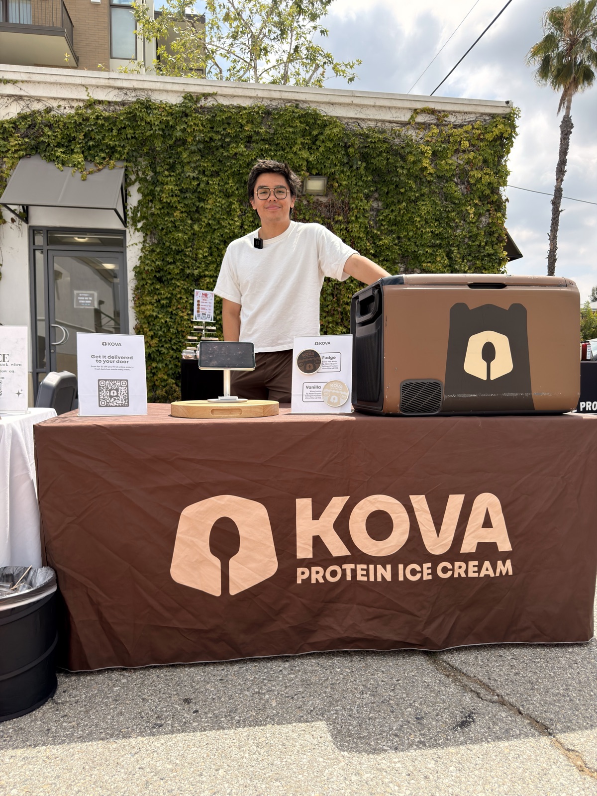 A Kova pop-up table: the full lockup printed across the tablecloth, the snout icon on the wrapped freezer, and printed flavor cards and a QR sign on the table.